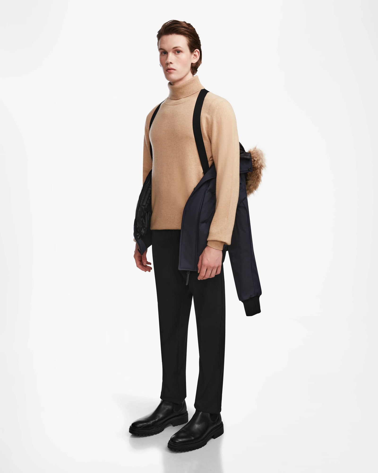 A young man stands against a white background wearing a beige turtleneck, black pants, and boots. He carries the Rudsak DALLAS - M122501 Navy puffer with a fur-trimmed hood over one shoulder by its straps.