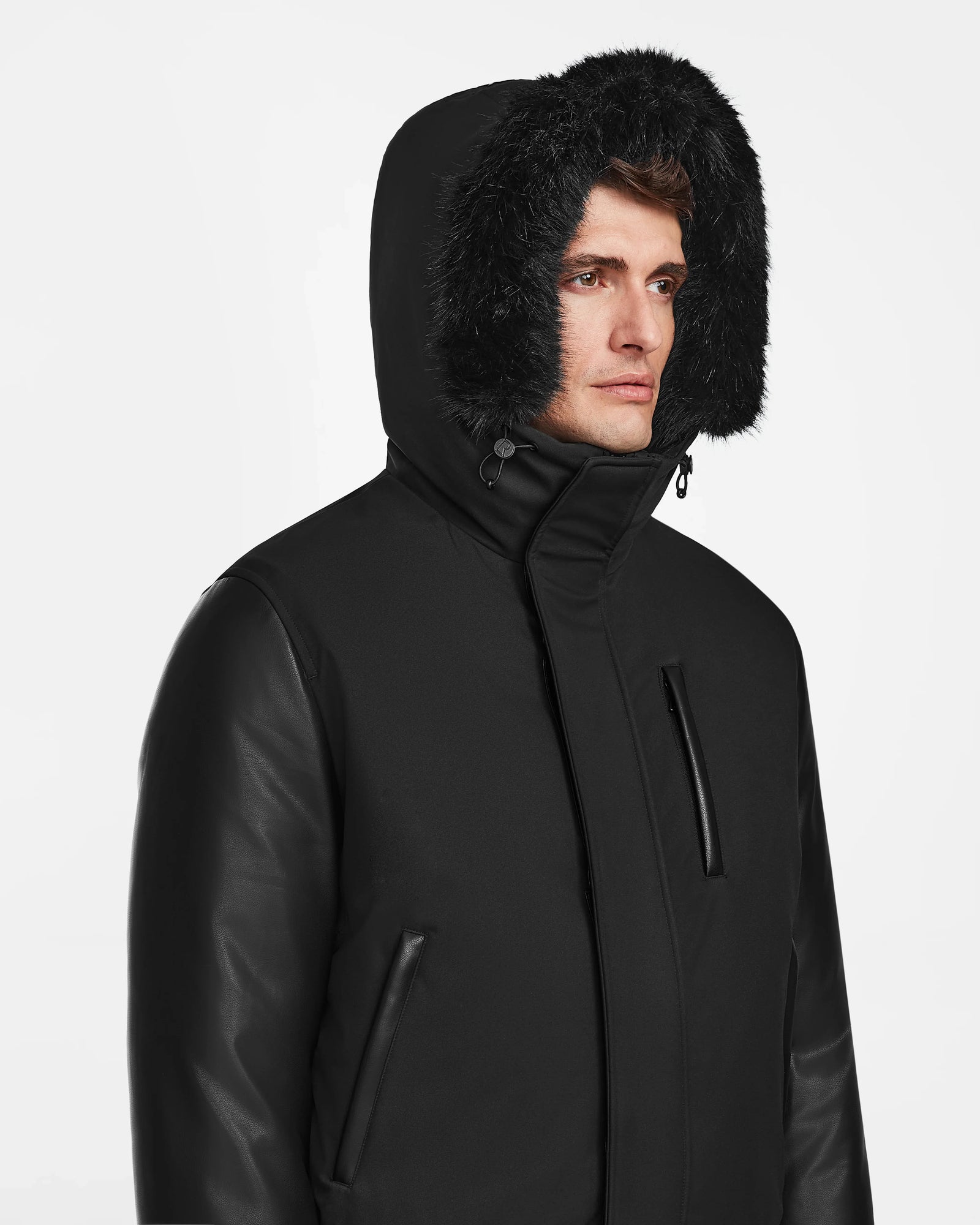 A man looks to the side wearing Rudsak’s DERON - M123517 BLACK bomber jacket, featuring a weather-resistant finish, faux fur-lined hood, zippered pockets, and leather sleeves, set against a plain white background.