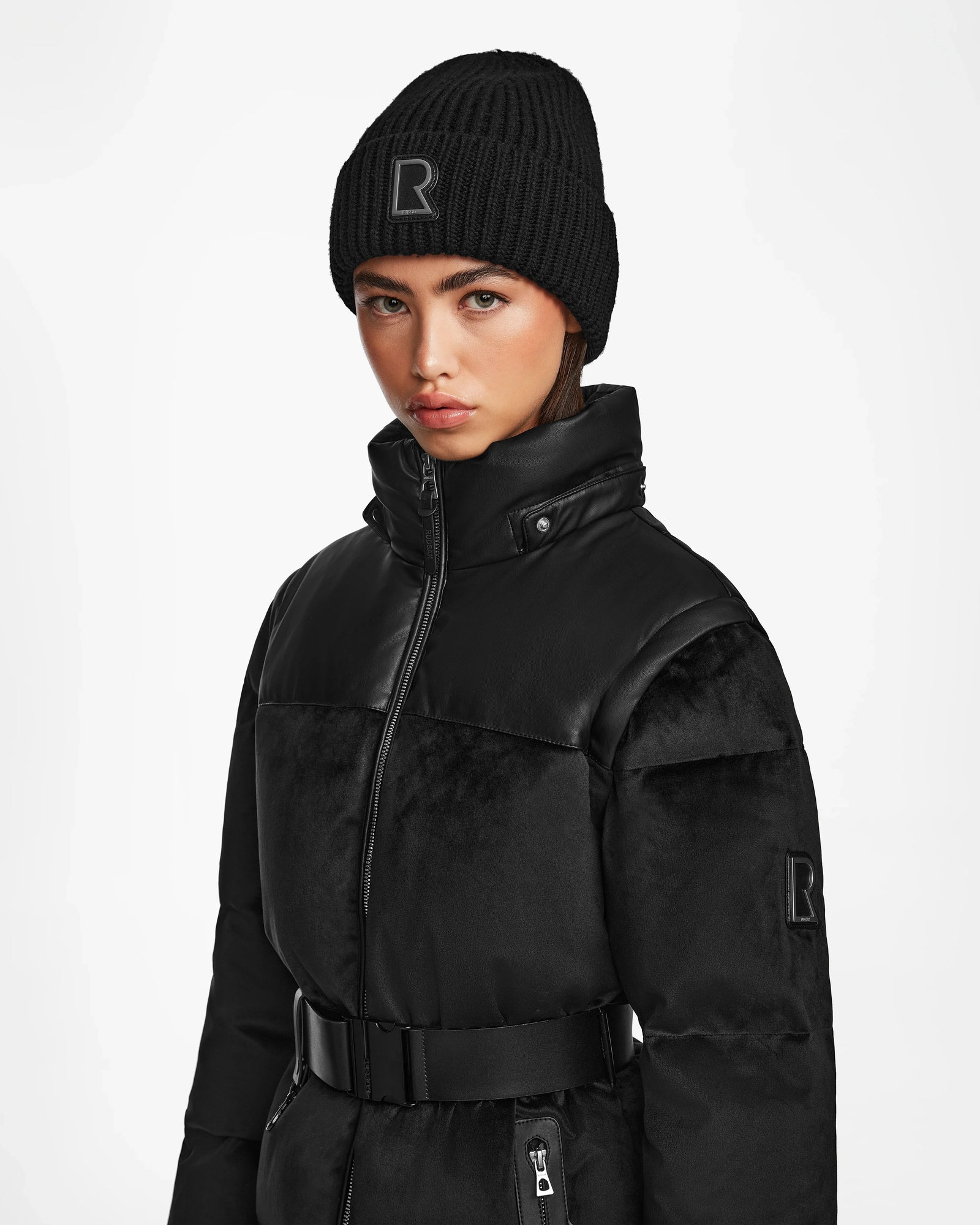 A woman wears a black knit beanie and the DONIA - L124563 Black women’s down puffer jacket with belt by Rudsak, both featuring an R patch, against a plain white background. The jacket doubles as a stylish convertible winter coat.