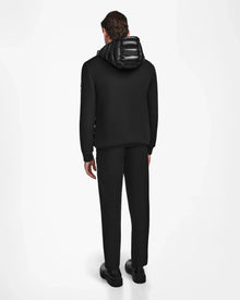 A person stands facing away from the camera, wearing the Rudsak DURIN S - M123518 Black mens hooded jacket with shiny hood and lightweight down insulation, paired with matching black pants and shoes against a plain white background.