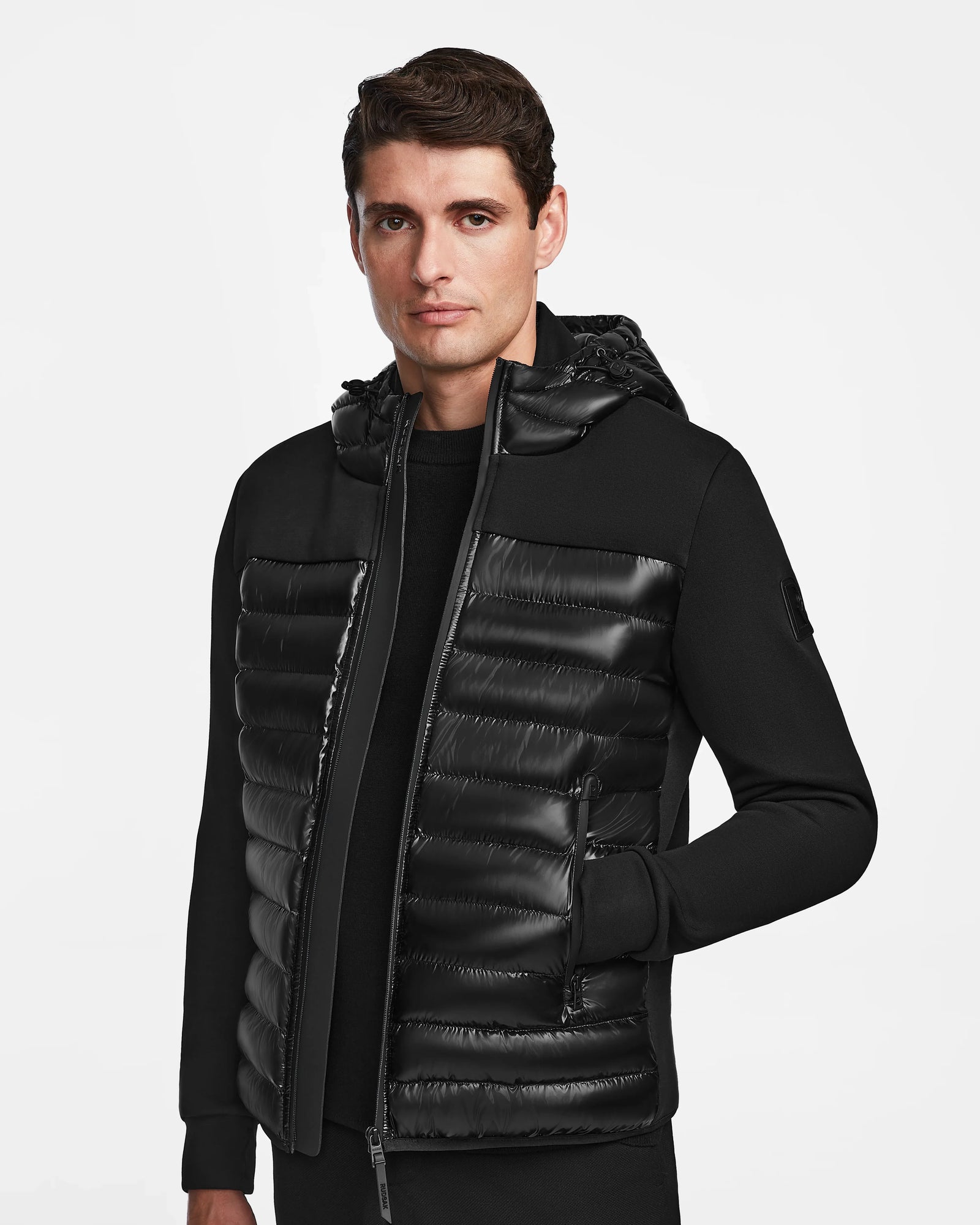 A man wears the Rudsak DURIN S - M123518 Black mens hooded jacket with lightweight down insulation, standing against a plain white background and looking slightly to the side with a neutral expression.
