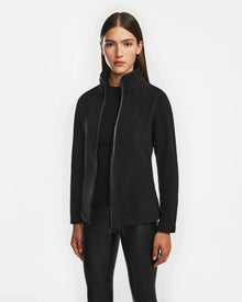 A woman with straight brown hair wears the Rudsak EDITH - 8524617 Black zip-up sweater over a black top and black leggings, standing against a plain white background.