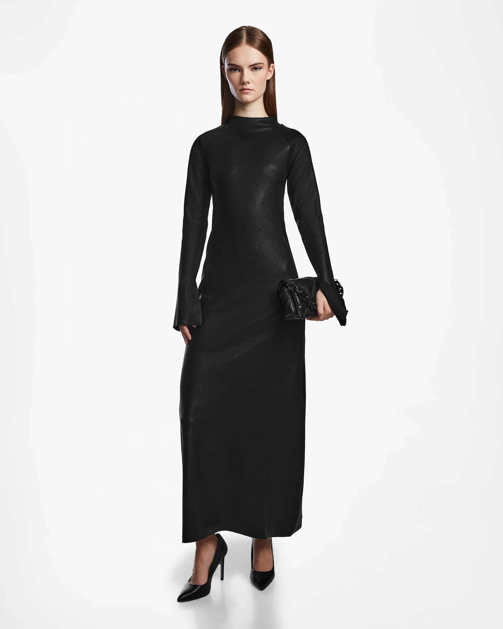 A woman with straight brown hair wears the Rudsak FAIZA - 8524579 Black maxi dress with long sleeves and black heels, holding a black clutch against a plain white background, highlighting contemporary style.