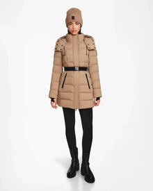 A woman stands facing forward against a plain white background, wearing the Rudsak FRIDA M - L124524 Sand mid-length down jacket along with a matching tan knit beanie, black leggings, and black ankle boots.