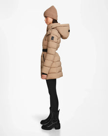 A woman wearing the Rudsak FRIDA M - L124524 Sand quilted down jacket with removable hood, a tan knit beanie, black leggings, and black boots stands in profile against a plain white background.