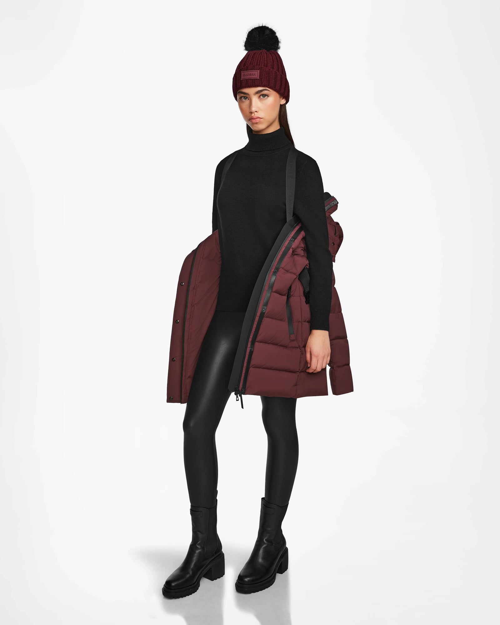 A woman wears a black turtleneck, leggings, boots, and Rudsaks FRIDA M - L124524 Wine mid-length down jacket draped over her arms with a matching maroon beanie featuring a black pom-pom, set against a plain white background.