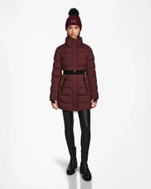 A woman models the Rudsak FRIDA M - L124524 Wine, a stylish womens mid-length down jacket with a removable hood, black belt, and matching beanie, paired with black leggings and boots against a white background.
