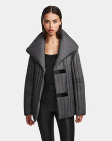 A woman with straight brown hair models the Rudsak INES T - 8124625 Grey, an oversized shiny dark gray quilted jacket with a wide collar, styled with a black top and pants against a plain white background.
