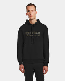 A man stands against a white background, wearing the Rudsak JARED - 6524533 Black mens hoodie, featuring RUDSAK CANADIAN HERITAGE printed in gold on the front.