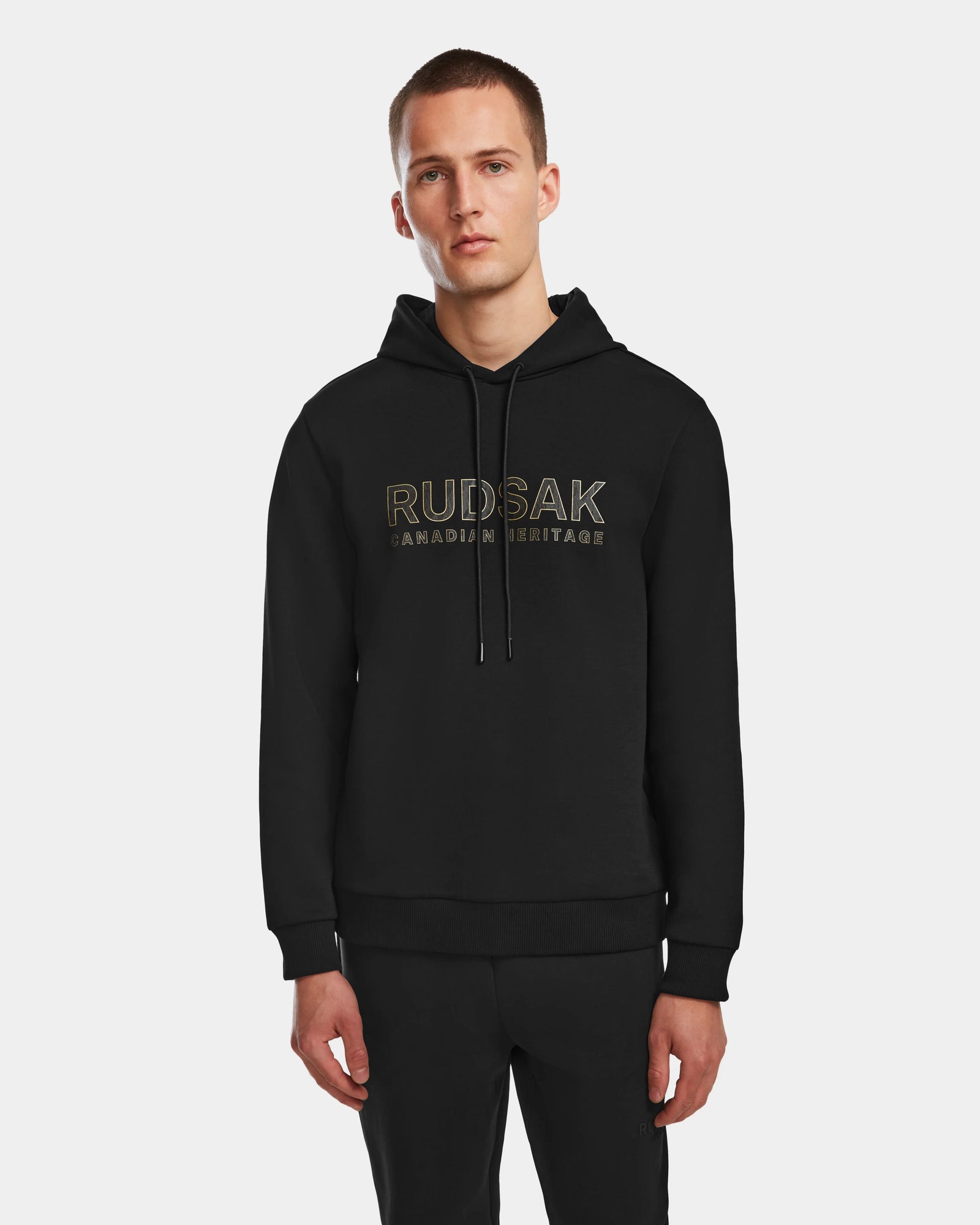 A man stands against a white background, wearing the Rudsak JARED - 6524533 Black mens hoodie, featuring RUDSAK CANADIAN HERITAGE printed in gold on the front.