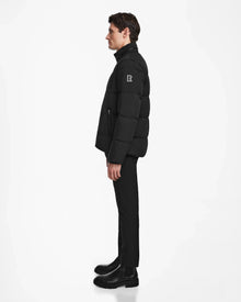 A man in profile wears the Rudsak KARLS - M124505 black mens puffer jacket with a logo on the upper sleeve, paired with black pants and boots, standing against a plain white background.