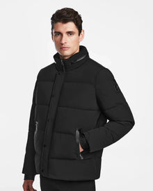 A man with dark hair wears the Rudsak KARLS - M124505 Black mens puffer jacket, featuring a high collar, quilted design, zippered pockets, and snap buttons. He stands against a plain white background, gazing slightly to the side.