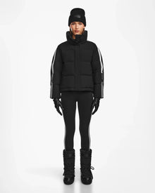 A person stands facing forward against a white background, wearing the Rudsak KIMIA - 8124716 Black ski jacket with white sleeve stripes, black leggings with white side stripes, black winter boots, gloves, and a black beanie.