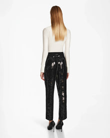 A woman with straight, long blonde hair stands facing away, wearing a white long-sleeve top, Rudsak LEAR - 8524553 Black sequin holiday trousers, and black high heels against a plain white background.