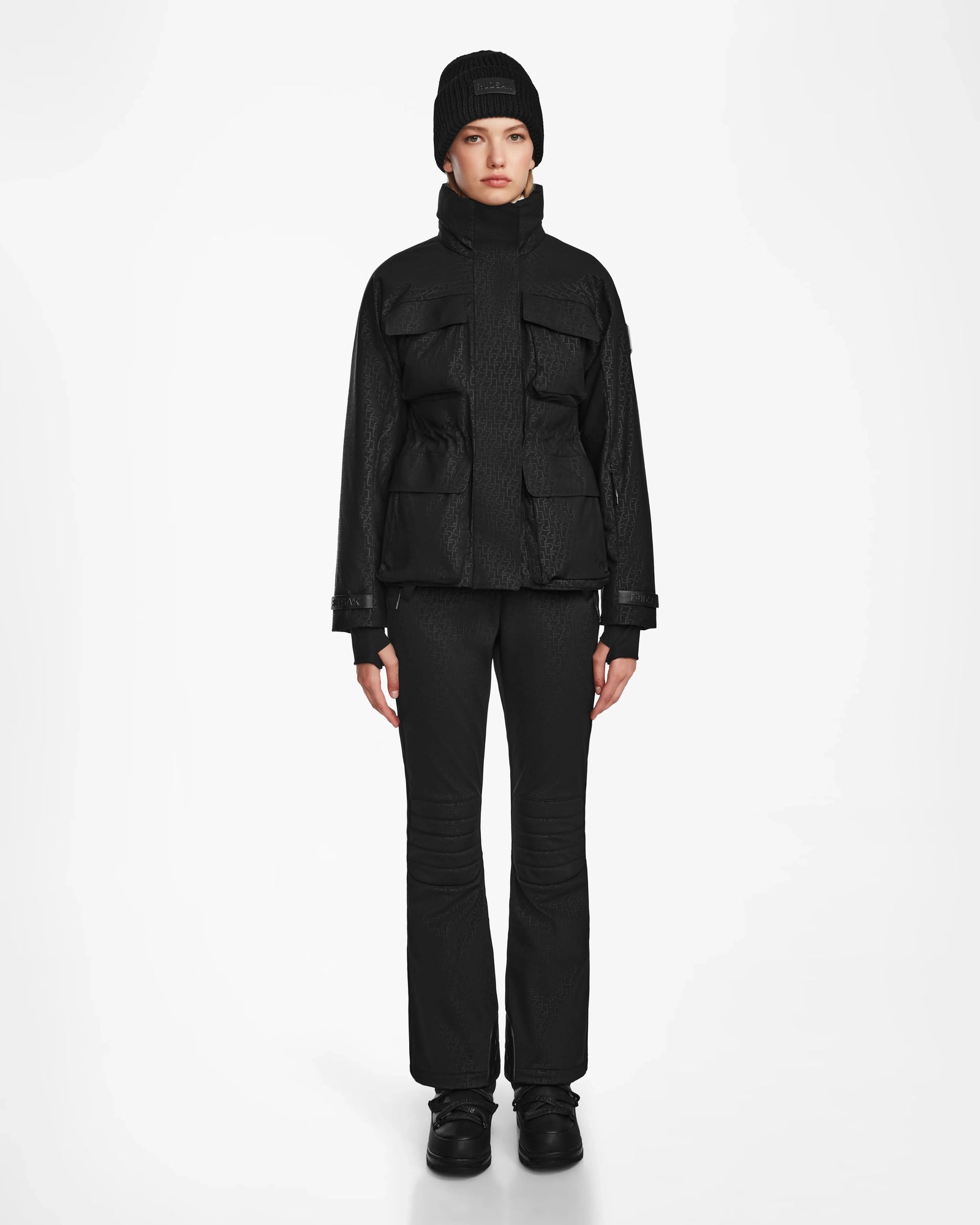 A person stands facing forward against a white background, wearing a Rudsak LUX MO - 8124570 Monogram Print Black ski jacket with large pockets, paired with black pants, shoes, and a black beanie for a sleek monochromatic look.