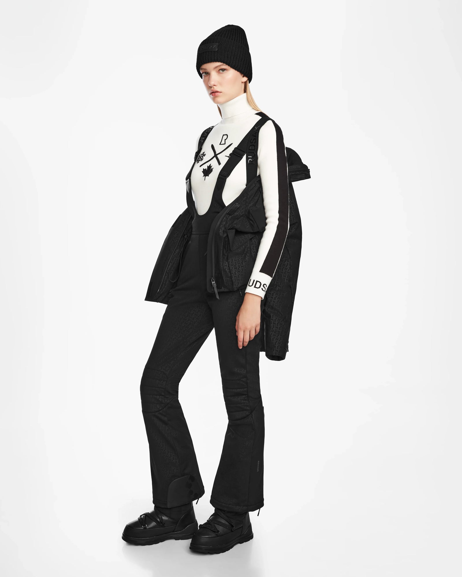 A woman stands against a white background wearing Rudsaks LUX MO - 8124570 Monogram Print Black ski jacket draped over her shoulders, styled with a black beanie, graphic white turtleneck, black overalls, and chunky black boots.