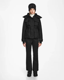 A woman models the Rudsak LUX MO - 8124570 Monogram Print Black ski jacket and matching pants against a white background, paired with black shoes, a white turtleneck, and a black cap. She gazes at the camera with a neutral expression.