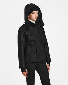 A person wearing the Rudsak LUX MO - 8124570 Monogram Print Black ski jacket, paired with black pants and a black beanie, stands against a plain white background, gazing slightly to the side with a neutral expression.