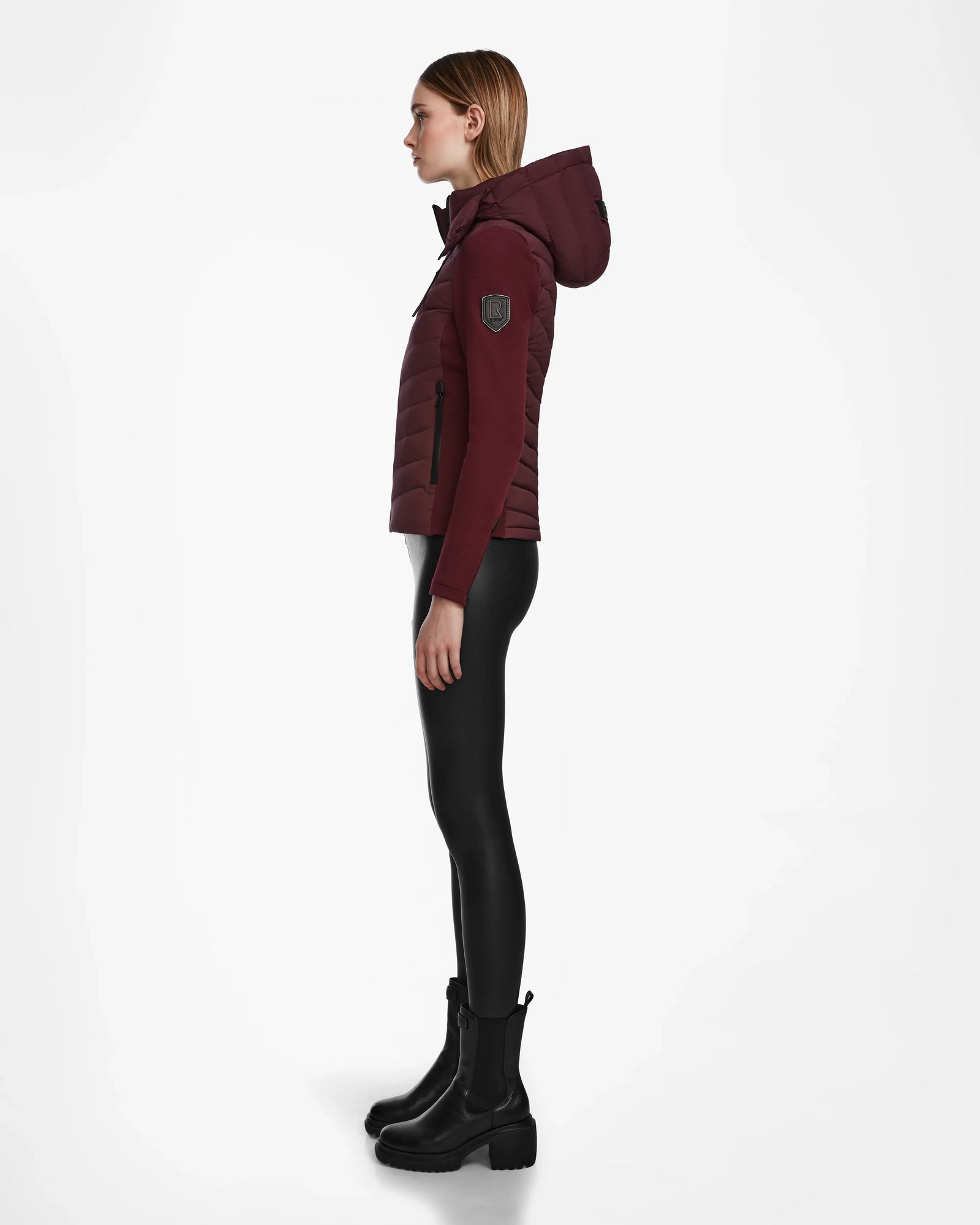 A woman stands in profile wearing the Rudsak MALYA M - L123547 Wine down quilted jacket, paired with black leggings and black boots against a plain white background.