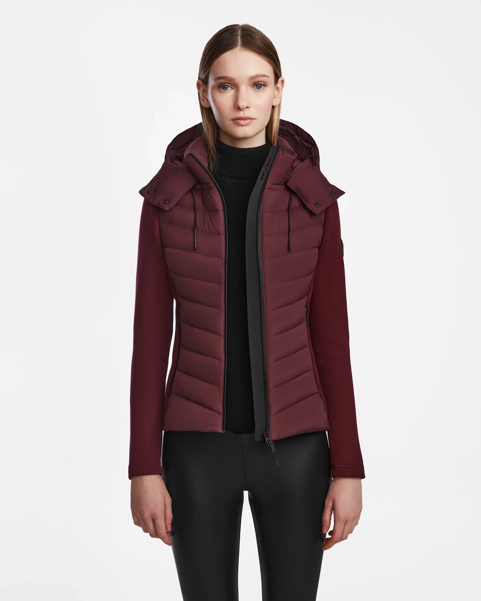 A woman with straight, light brown hair models the Rudsak MALYA M - L123547 Wine down quilted jacket over a black top and pants, standing against a plain white background.