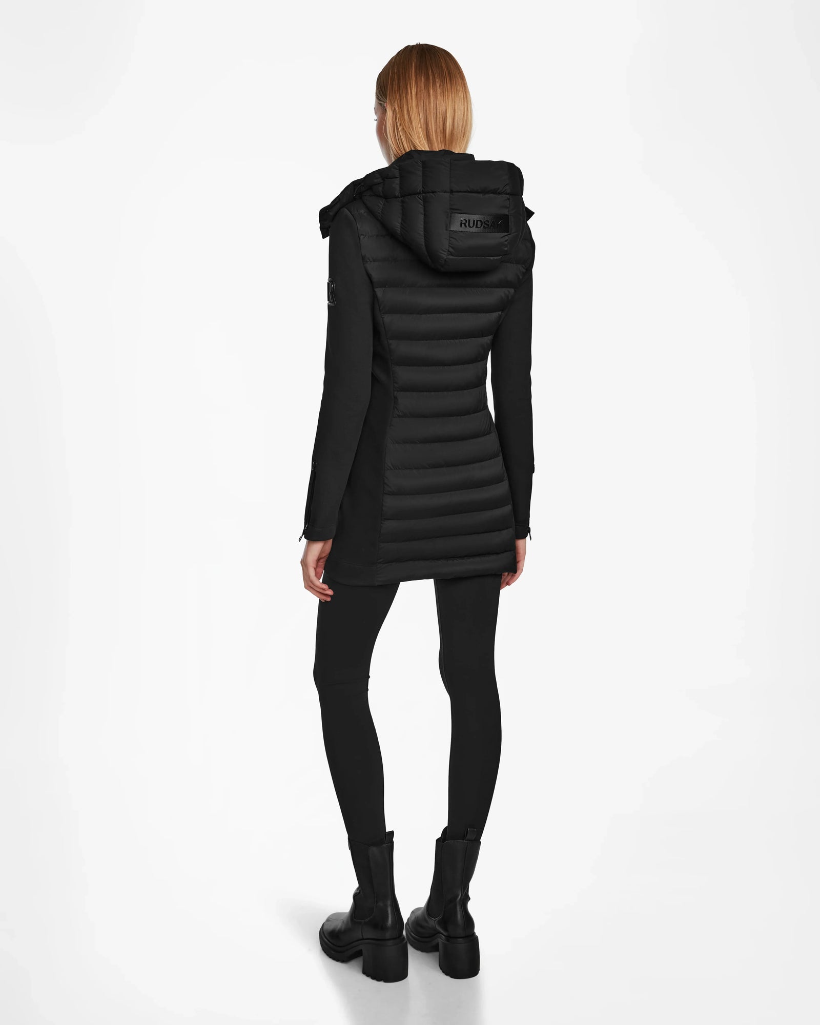 A person with straight, shoulder-length hair wears the RUDSAK MERINA - 8122546 BLACK quilted down jacket, black leggings, and boots, standing with their back to the camera against a white background.