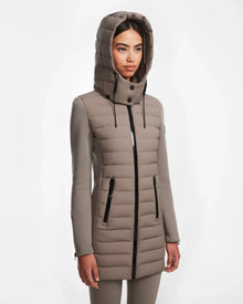 A woman with long dark hair and a neutral expression wears the Rudsak MERINA M - 8124030 Cement, a taupe hooded quilted down jacket with zippered pockets, standing against a plain white background.