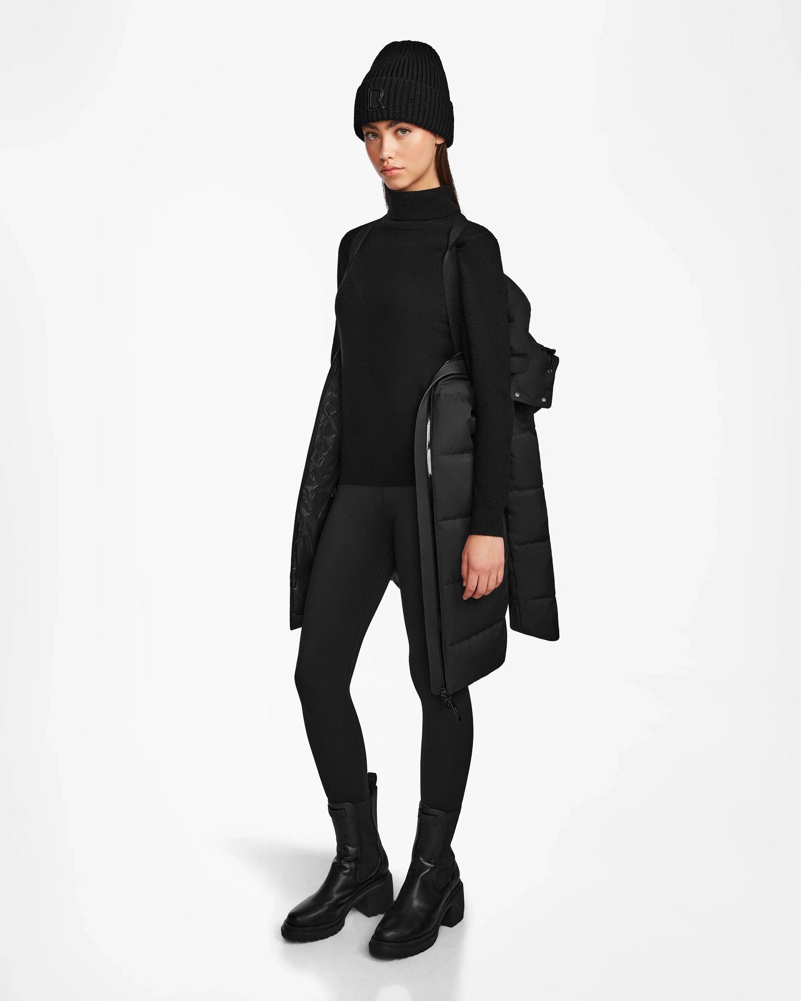A woman stands against a white background wearing the Rudsak MIRIAM - L122523 BLACK womens down puffer coat with a weather resistant shell draped over one shoulder—an ideal addition to any winter wardrobe.