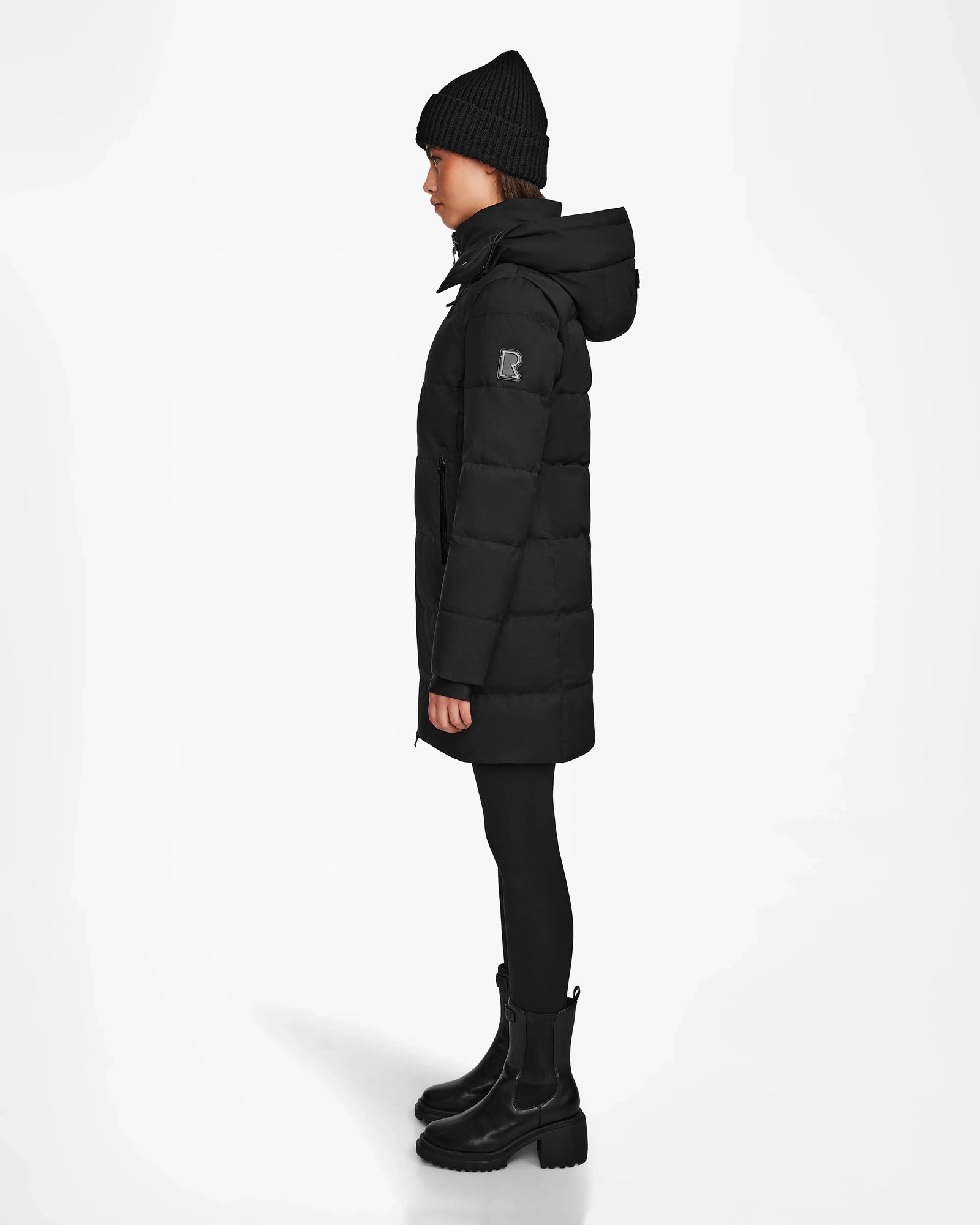 Wearing the Rudsak MIRIAM - L122523 BLACK weather-resistant puffer coat with black beanie, pants, and boots, this look in profile against a white background is ideal for any winter wardrobe.