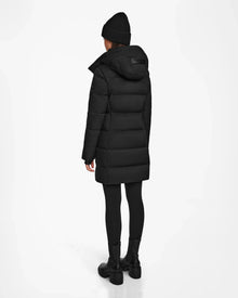 A person wears the Rudsak MIRIAM - L122523 BLACK womens down puffer coat with black pants, boots, and a knit beanie, standing against a white background—a chic winter staple.