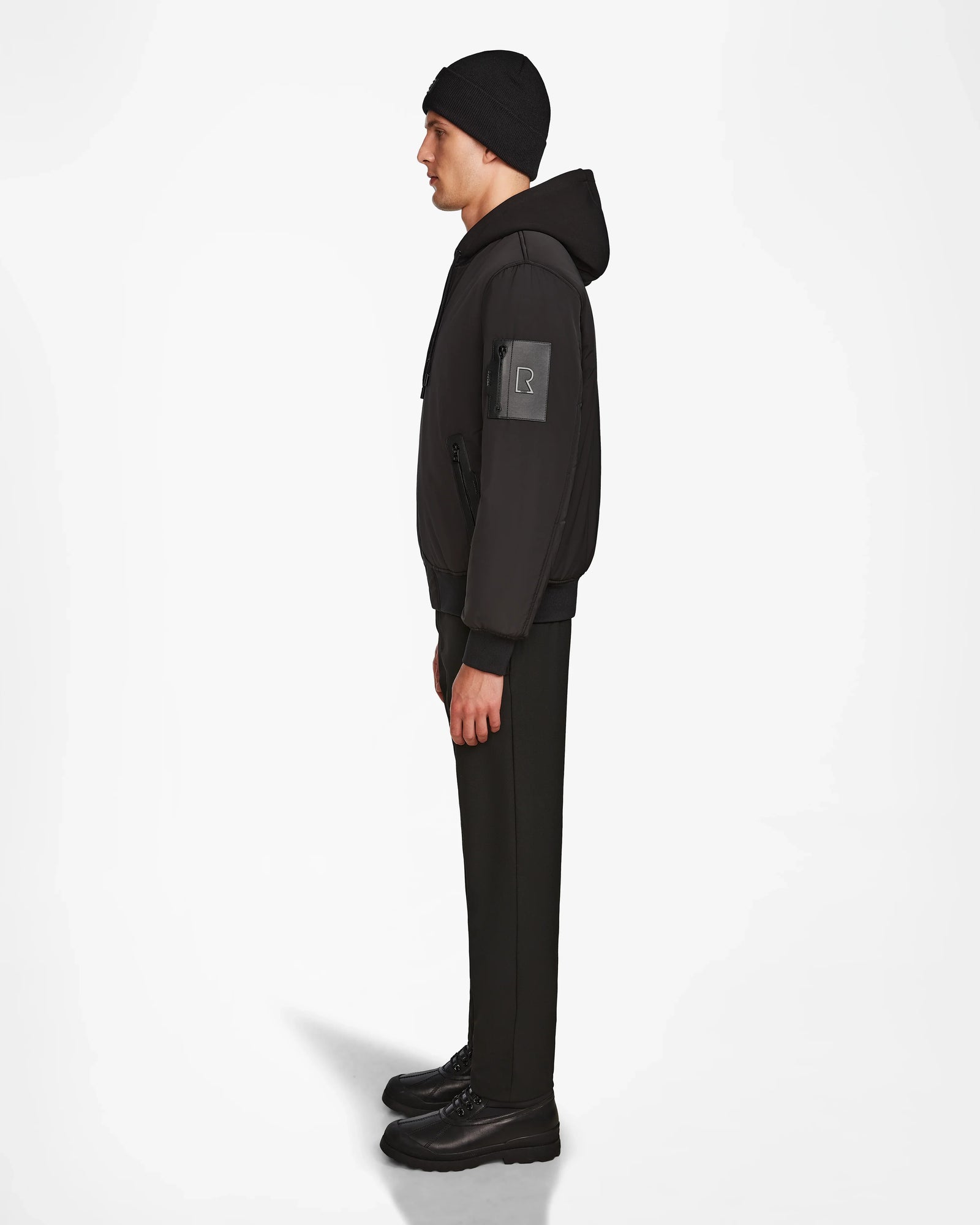 A man in profile faces left, wearing Rudsaks NEVEN - M124520 Black mens bomber jacket with a sleeve patch made from recycled materials, paired with a black beanie, pants, and shoes against a plain white background.