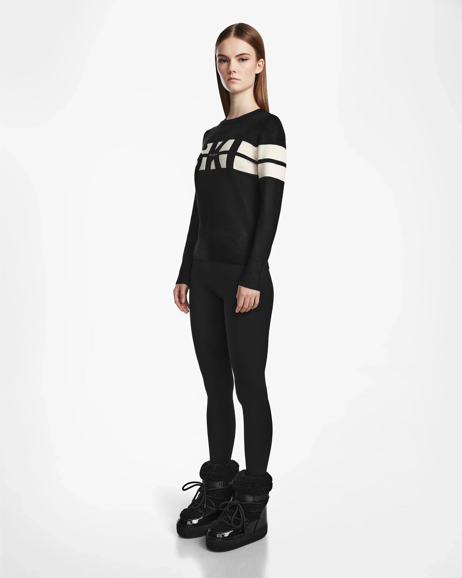 A woman with straight brown hair stands against a white background wearing Rudsaks ODESSA - 8523540 Black crewneck sweater with two white stripes and bold lettering, paired with black leggings and shiny black winter boots.