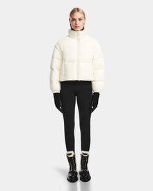 A woman stands facing forward wearing the Rudsak OPRAH - 8124708 Iceland womens cropped down puffer jacket with black gloves, leggings, and winter boots with white trim against a plain light gray background.