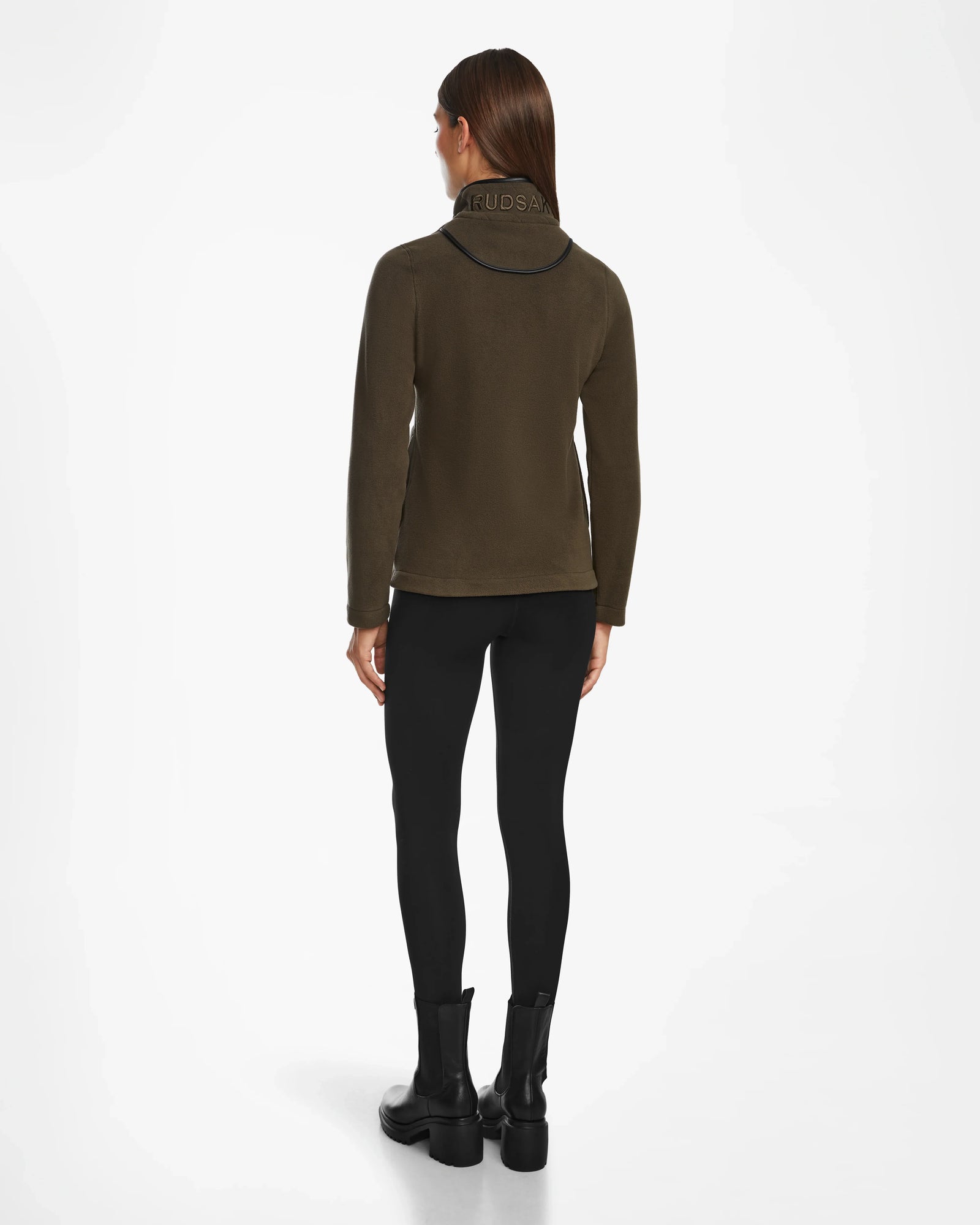 A woman faces away, wearing the Rudsak ORCHID - 8524616 Army womens fitted half-zip pullover with vegan leather trims, black leggings, and ankle boots against a plain white background.