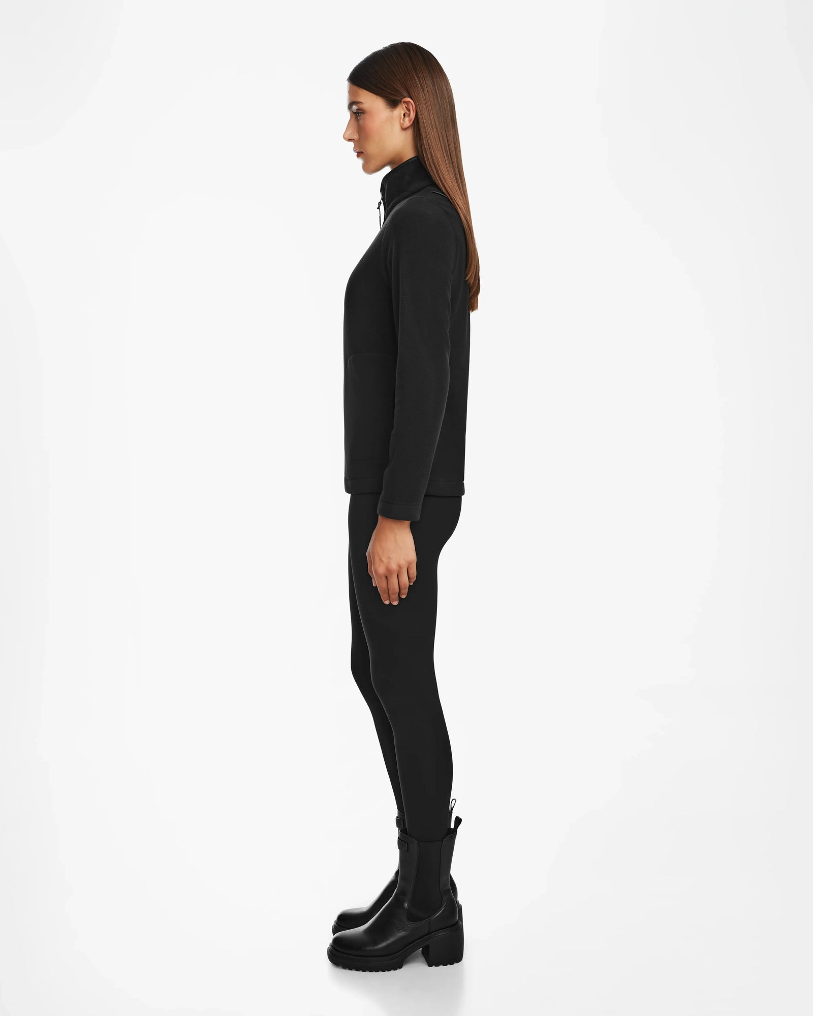 A woman with straight brown hair stands in profile, wearing the Rudsak ORCHID - 8524616 Black zip-up jacket with vegan leather trims, black leggings, and black boots against a plain white background.