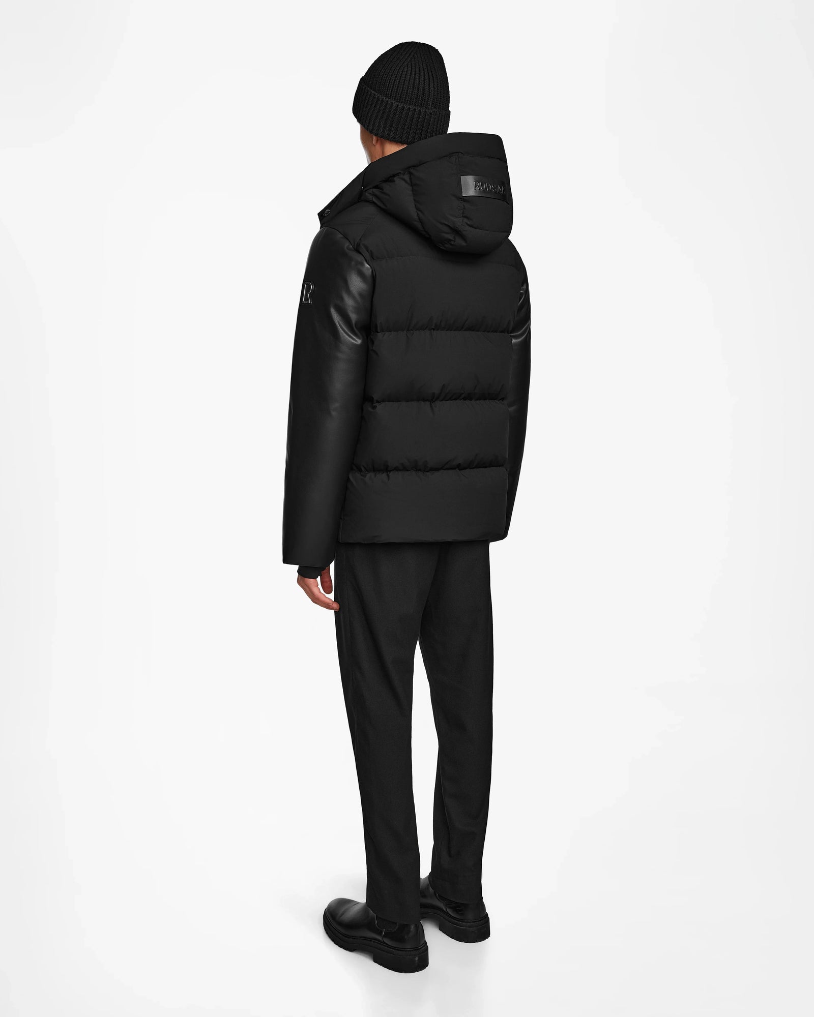 A person stands facing away, wearing the Rudsak RONNY M - M124513 Black hooded puffer jacket made from recycled materials, paired with black pants and shoes against a plain white background.