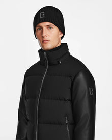 A man stands against a white background wearing the Rudsak RONNY M - M124513 Black quilted down jacket with leather sleeves and matching black knit beanie, both featuring the signature R logo—an essential modern winter look.
