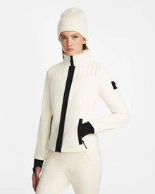 A woman wearing the Rudsak ROSALIE - 8124784 Iceland women’s ski jacket with RECCO rescue tech, white pants, and a white knit beanie stands with one hand in her pocket against a plain white background.