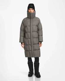 A person wearing the Rudsak SASH - 8124536 Light Grey women’s long down puffer coat, black leggings, winter boots, and a black beanie faces forward against a plain white background.