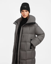 A woman models the Rudsak SASH - 8124536 Light Grey womens long down puffer coat and a black knit beanie, standing against a plain white background and highlighting its stylish winter design with RDS-certified down fill.