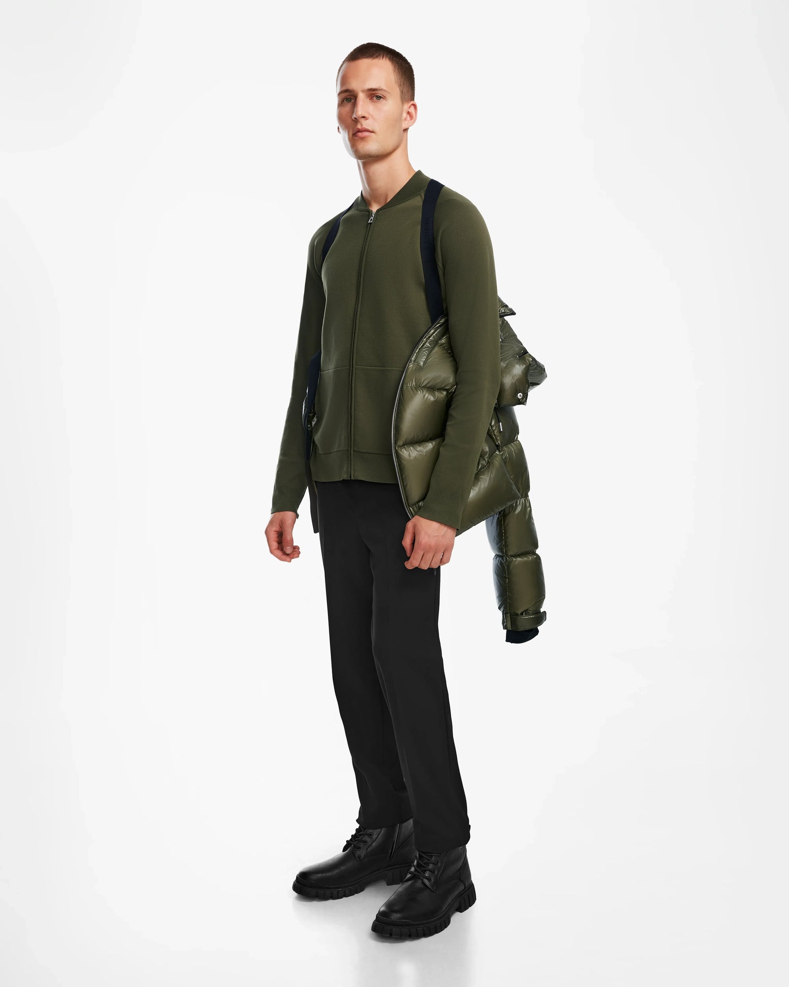 A man stands against a white background, wearing the Rudsak VINCE S - 6124560 Loden Green jacket, black pants, and black boots. He holds a mens down puffer over his shoulder and looks straight ahead.
