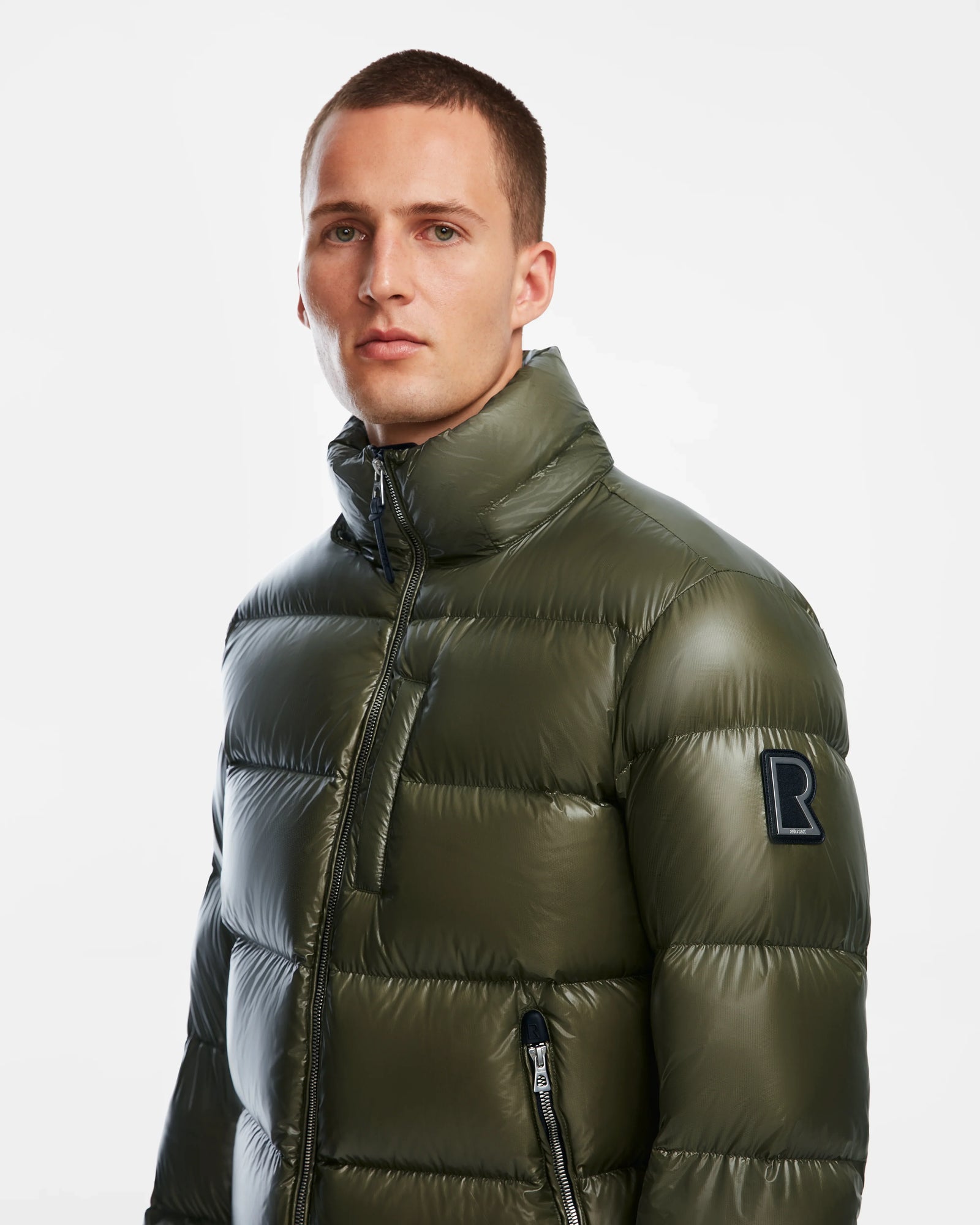 A man wearing a Rudsak VINCE S - 6124560 Loden Green down puffer jacket with a high collar and black R patch on the sleeve stands against a plain white background.