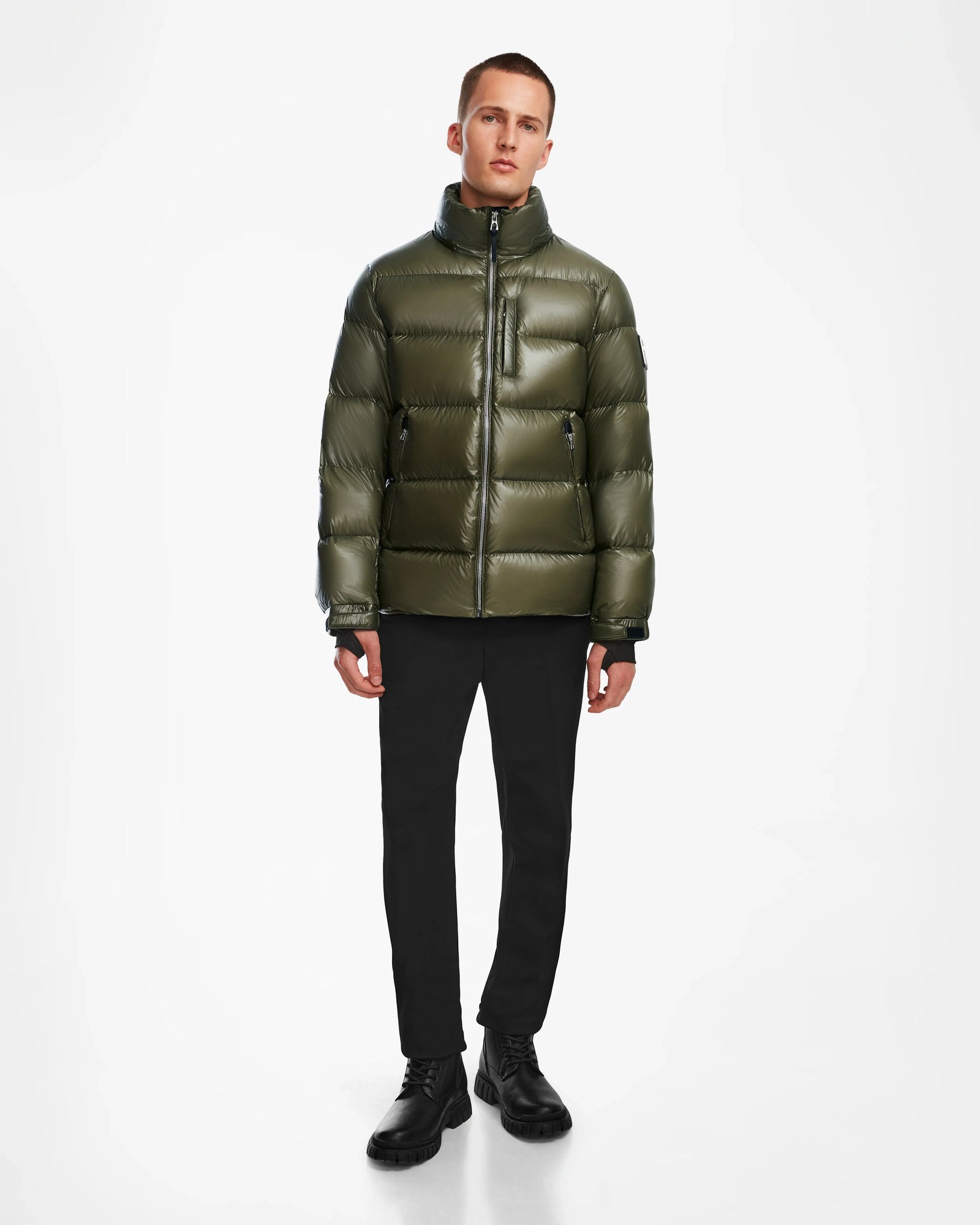 A person stands against a white background wearing the Rudsak VINCE S - 6124560 Loden Green mens down puffer jacket, which has a shiny weather-resistant finish, high collar, front zipper, zippered chest pocket, black pants, and black shoes.
