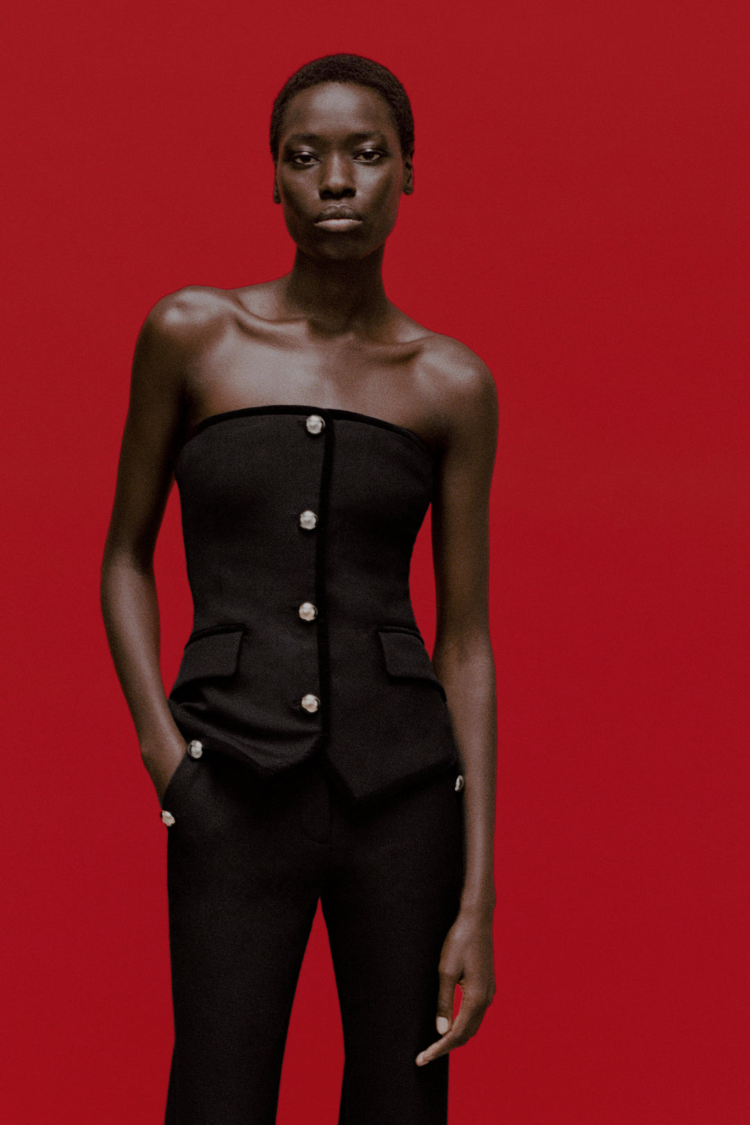 Black strapless bustier top with pearl button front closure and side pockets, worn with matching trousers.