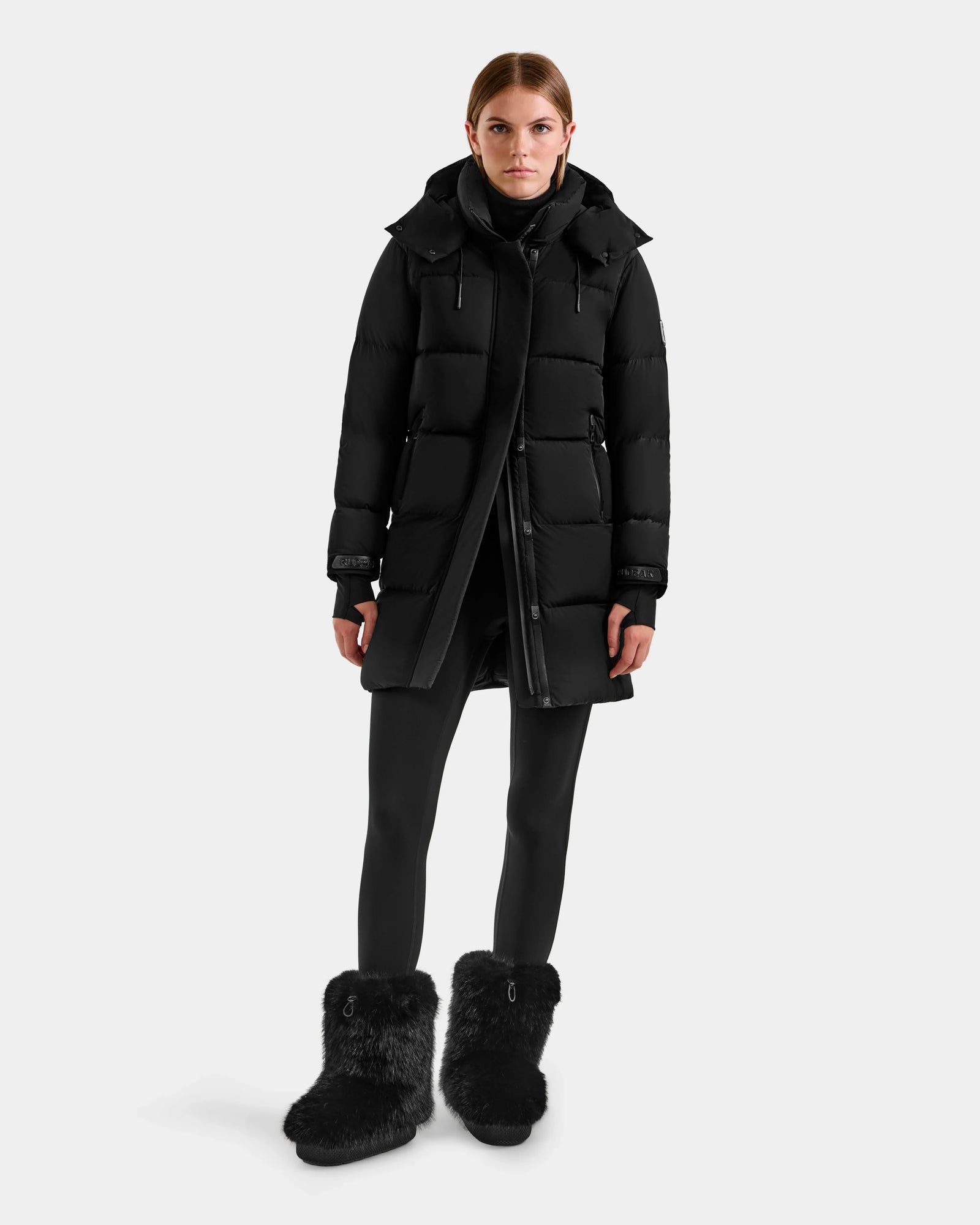 A person stands facing forward in the Rudsak ALESSANDRA M - L125552 Black women's down puffer long winter coat, with black leggings and furry boots against a plain light gray background.