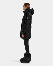 A person stands in profile against a plain light background wearing the Rudsak ALESSANDRA M - L125552 Black women’s down puffer with a hood, paired with black leggings and black furry boots.
