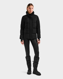 A woman stands against a white background, wearing the Rudsak ALESSANDRA S-M - L125551 Black down puffer jacket with shiny leggings and black boots. Facing forward with arms relaxed, she showcases this cold-weather essential.