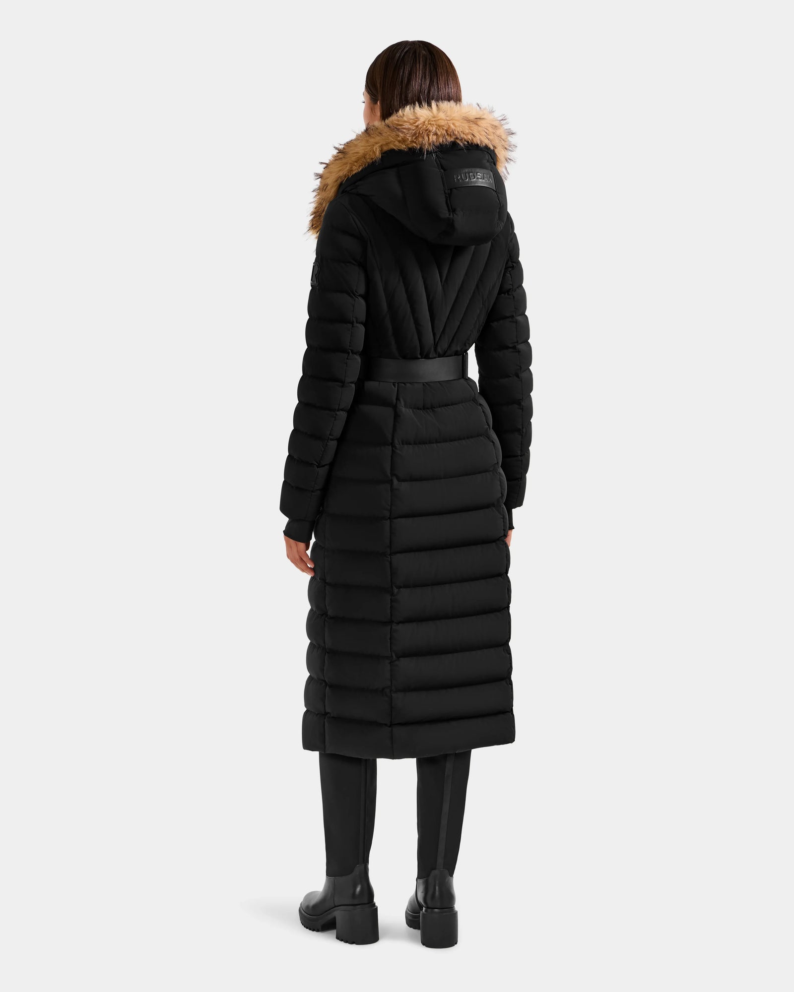 A person wearing the Rudsak ALISA L - L125574 Black long, quilted puffer coat with faux-fur trim hood, black pants, and boots stands against a light gray background, shown from the back.