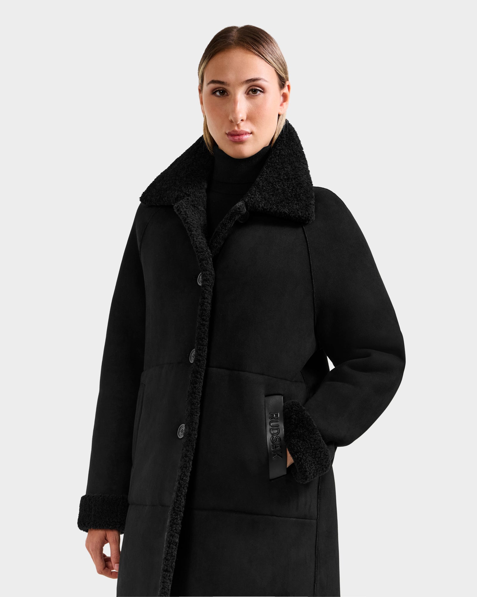 A woman stands facing forward in the Rudsak ANGILICA - L125600 Black coat, featuring a textured collar and cuffs, buttoned front, and black turtleneck underneath, with one hand in her pocket against a plain light gray background.