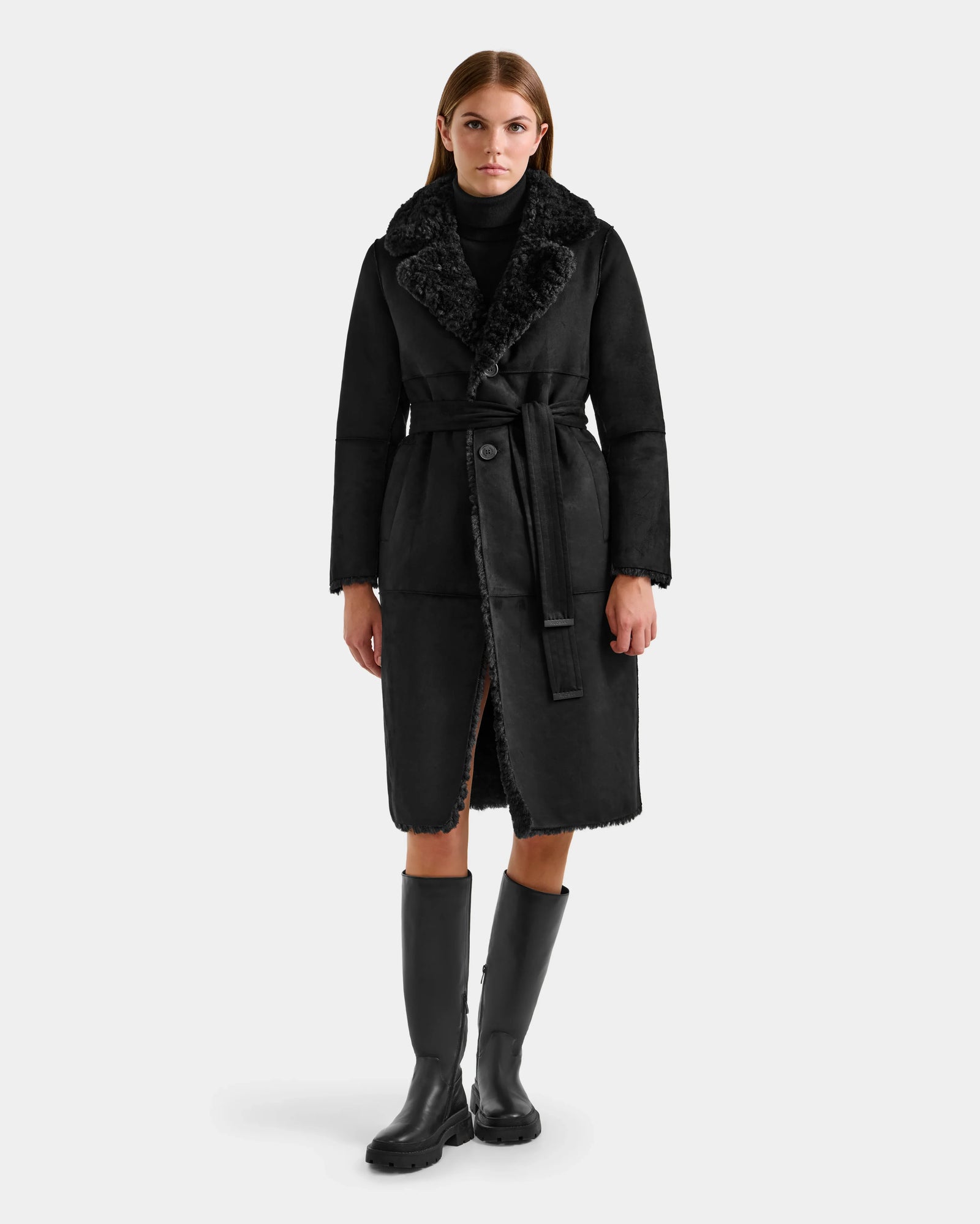 A woman faces forward against a light gray background, wearing the Rudsak ANNABELLE FSU - L125512 Black faux suede trench coat with a tie belt, paired with a black turtleneck and knee-high black boots.
