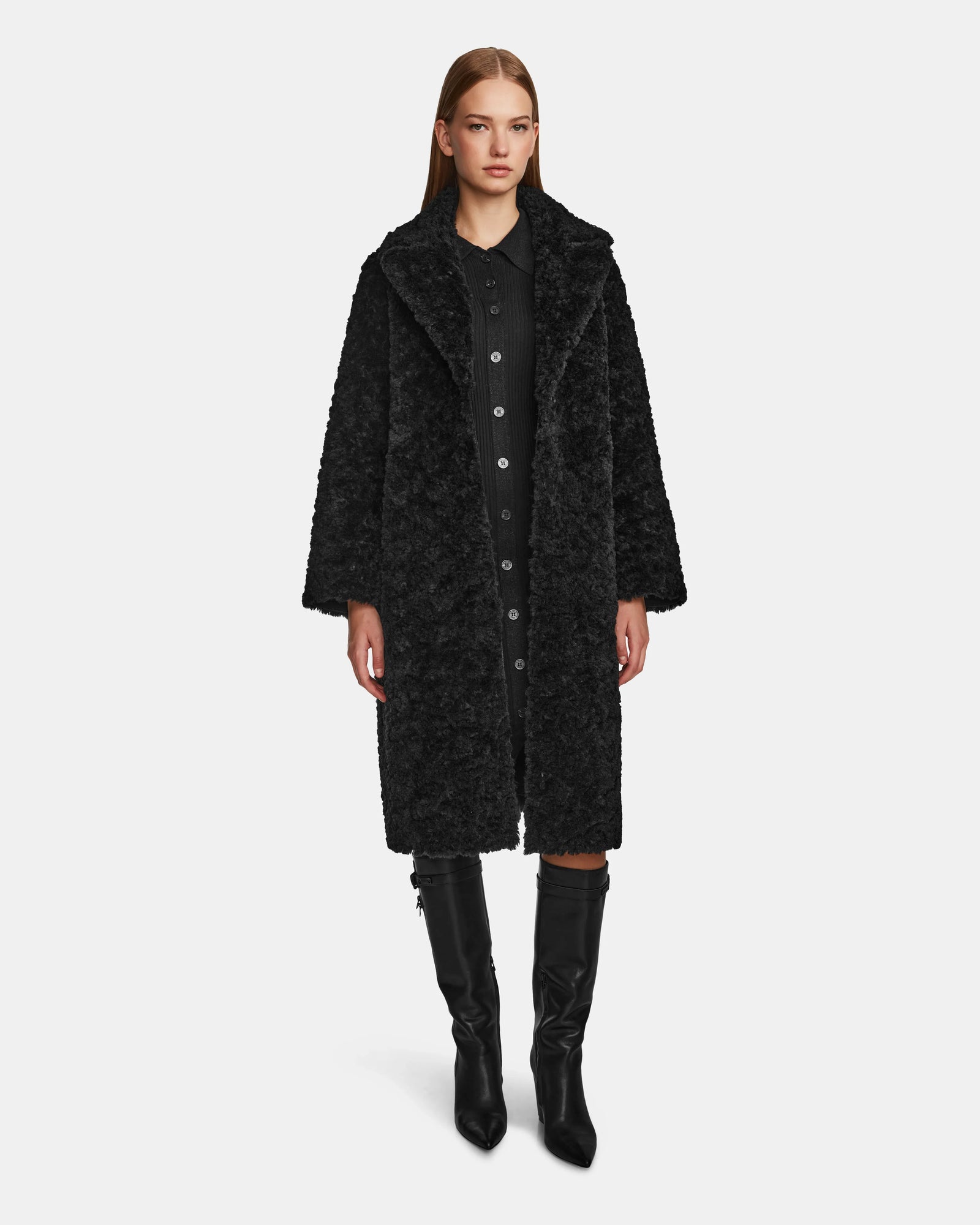 A woman stands facing forward against a plain white background, wearing the Rudsak ANNABELLE - L124544 Black textured faux fur coat over a buttoned black dress and black knee-high heeled boots.
